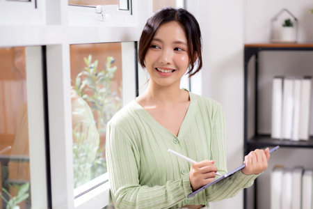 Young asian woman using tablet writing with inspiration and comfortable by window in living room at home office, cheerful woman standing working on digital tablet in workspace, business and lifestyle.の写真素材