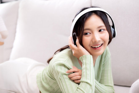Happy young asian woman wearing headphones listening music while lying on sofa with fun and enjoy in the living room at home, happiness female with entertainment, one person, lifestyle concept.の写真素材