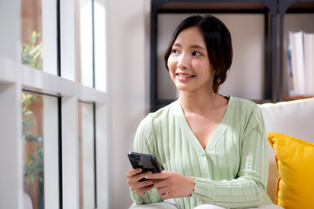Young asian businesswoman looking smartphone smiling and relax sitting on sofa in living room at home, cheerful woman watching smart phone sitting on couch in home, lifestyle and communication.の写真素材