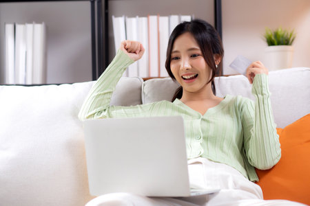 Young asian woman sitting on sofa using laptop computer shopping online with credit card buying to internet while cheerful, female payment with business, purchase and payment, business and finance.の写真素材