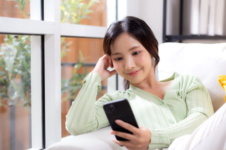 Young asian businesswoman looking smartphone smiling and relax sitting on sofa in living room at home, cheerful woman watching smart phone sitting on couch in home, lifestyle and communication.の写真素材