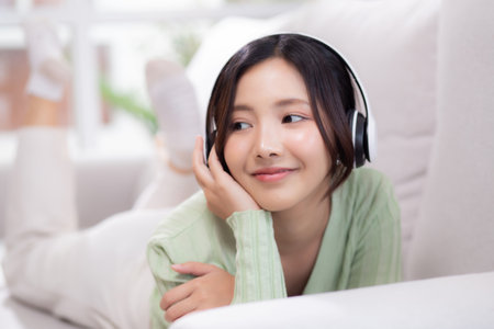 Happy young asian woman wearing headphones listening music while lying on sofa with fun and enjoy in the living room at home, happiness female with entertainment, one person, lifestyle concept.の写真素材