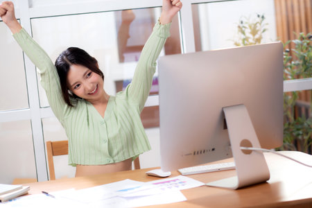 Young asian business woman using desktop computer for success in modern home office, businesswoman working with desktop computer while winning and satisfied in living room at home, business concept.の写真素材