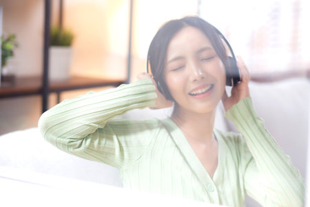 Happy young asian woman wearing headphones listening music while sitting on sofa with fun and enjoy in the living room at home, happiness female with entertainment, one person, lifestyle concept.の写真素材