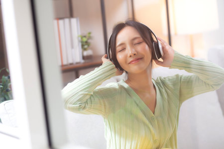 Happy young asian woman wearing headphones listening music while sitting on sofa with fun and enjoy in the living room at home, happiness female with entertainment, one person, lifestyle concept.の写真素材