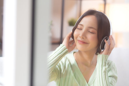Happy young asian woman wearing headphones listening music while sitting on sofa with fun and enjoy in the living room at home, happiness female with entertainment, one person, lifestyle concept.の写真素材