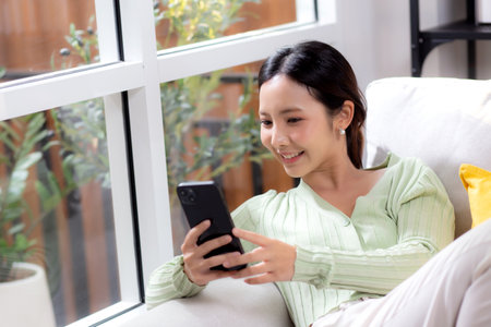 Young asian businesswoman looking smartphone smiling and relax sitting on sofa in living room at home, cheerful woman watching smart phone sitting on couch in home, lifestyle and communication.の写真素材