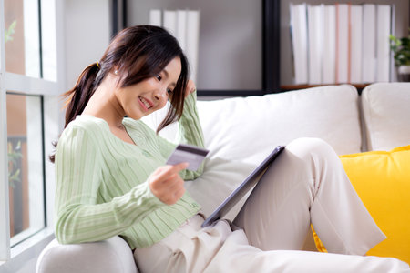 Young asian woman sitting on sofa using tablet computer shopping online with credit card buying to internet while thoughtful, female payment with business, purchase and payment, business and finance.の写真素材