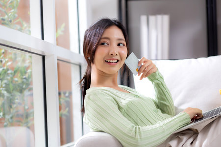 Young asian woman sitting on sofa using laptop computer shopping online with credit card buying to internet while thoughtful, female payment with business, purchase and payment, business and finance.の写真素材