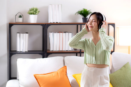 Happy young asian woman wearing headphones listening music while standing dancing with fun and enjoy in the living room at home, happiness female with entertainment, one person, lifestyle concept.の写真素材