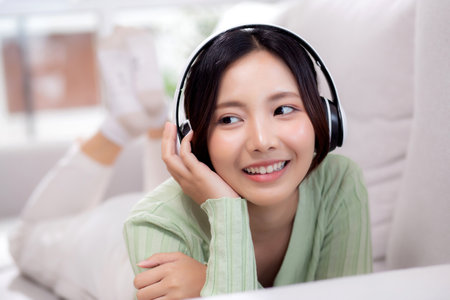 Happy young asian woman wearing headphones listening music while lying on sofa with fun and enjoy in the living room at home, happiness female with entertainment, one person, lifestyle concept.の写真素材