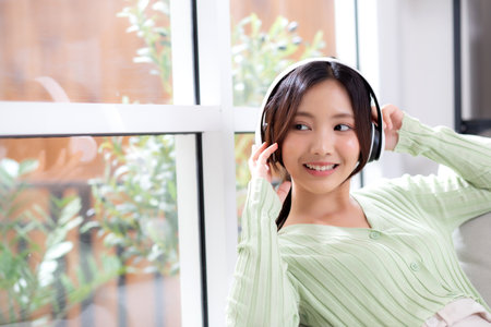 Happy young asian woman wearing headphones listening music while sitting on sofa with fun and enjoy in the living room at home, happiness female with entertainment, one person, lifestyle concept.の写真素材