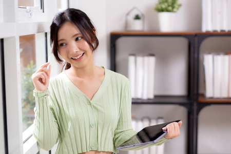 Young asian woman using tablet writing with inspiration and while thoughtful by window in living room at home office, cheerful woman standing working on digital tablet in workspace, business concept.の写真素材
