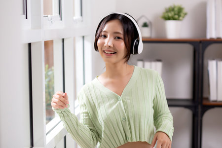 Portrait happy young woman wearing headphones listening music while standing with fun and enjoy in the living room at home, happiness female with entertainment, one person, lifestyle concept.の写真素材