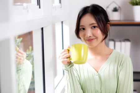 Portrait young asian woman relax and enjoyment with drinking coffee by the window in living room at home, woman smiling with happiness and comfortable drinking beverage at home, lifestyle and leisure.の写真素材