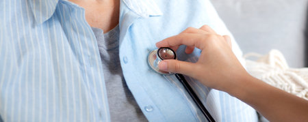 Caregiver young nurse woman examining elderly with stethoscope for checkup sitting on sofa in living room at home, nurse checkup patient about cardiac, patient and insurance, medical and health.の写真素材