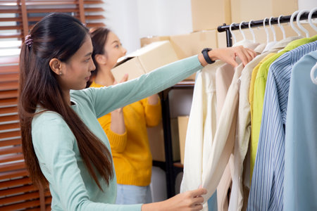 Two young asian woman arranging clothes in store for preparation delivery, seller choice clothes, seller choice clothes for retail online, merchandise for service with delivery, small business.の写真素材