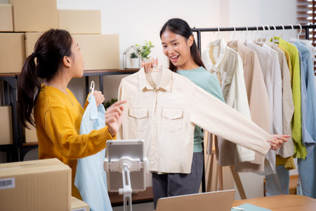 Two young asian woman live online clothing store while showing clothing with tablet on social at home, entrepreneur streaming for selling online clothing and presenting, online seller.の写真素材