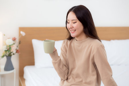Young asian woman holding and drinking coffee in morning in bedroom at home for relaxation, woman drinking beverage with pleasure and freshness for health, relax and cozy, lifestyle concept.の写真素材