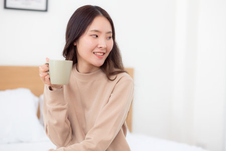 Young asian woman holding and drinking coffee in morning in bedroom at home for relaxation, woman drinking beverage with pleasure and freshness for health, relax and cozy, lifestyle concept.の写真素材