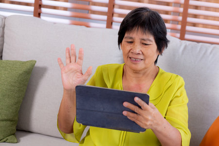 Senior asian woman waving during a video call on digital tablet sitting on sofa in living room at home, elderly video call online on tablet with distant sitting on couch, retirement and technology.の写真素材