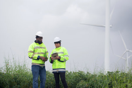 Two engineer check and examining wind turbines in wind farm, renewable energy and teamwork in a sustainable environment, inspection power electric with document and tablet, industry and eco.の写真素材