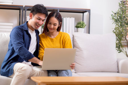 Young asian couple looking on laptop for relax with cozy and contentment sitting on sofa in living room at home, couple leisure watching laptop on couch, relationship and bonding, lifestyle concept.の写真素材