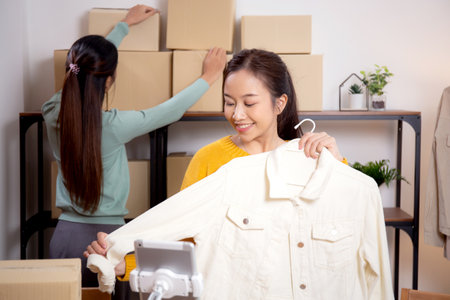 Two young asian woman live online clothing store while showing clothing with tablet on social at home, entrepreneur streaming for selling online clothing and presenting, online seller.の写真素材