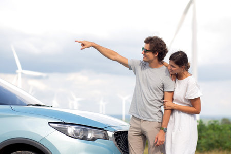 Young couple standing by electric car while wind turbines, renewable and innovation, windmill with resource sustainable power, renewable and environment with efficiency, power with eco.の写真素材