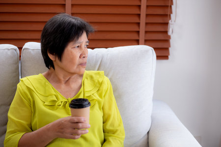 Happiness senior woman sitting on sofa and holding a coffee cup drinking looking out a window in living room at home, elderly drinking beverage for relax, lifestyle and cozy, domestic life.の写真素材