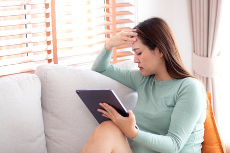 Young asian business woman using tablet computer stress and worried sitting on sofa in living room at home office, businesswoman working on tablet while concerned and tire, business and lifestyle.の写真素材