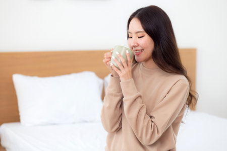 Young asian woman holding and drinking coffee in morning in bedroom at home for relaxation, woman drinking beverage with pleasure and freshness for health, relax and cozy, lifestyle concept.の写真素材