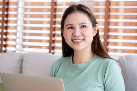 Portrait young asian business woman sitting on sofa using laptop computer in living room at home, businesswoman sitting on couch working on notebook with comfortable, business and freelance.の写真素材