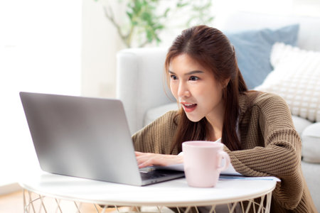 Young asian woman using laptop with surprise in living room at home, woman relaxation with notebook computer or working from home with shock, business browsing email, business and freelance.の写真素材