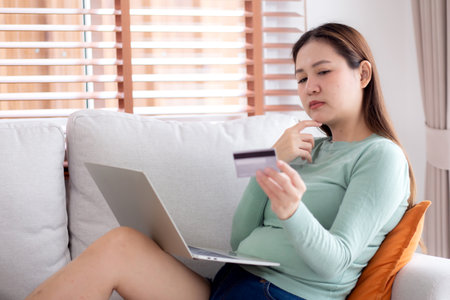 Young asian woman sitting on sofa using laptop computer shopping online with credit card buying to internet while thoughtful, female payment with business, purchase and payment, business and finance.の写真素材