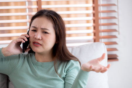 Asian young woman stress and worried sitting on sofa while speaking on smart phone in living room at home, woman unhappy and concerned talking on smartphone, lifestyle and communication.の写真素材
