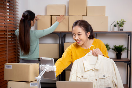 Two young asian woman live online clothing store while showing clothing with tablet and laptop on social at home, entrepreneur streaming for selling online clothing and presenting, online seller.の写真素材