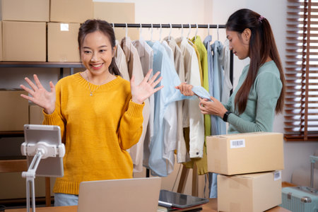 Two young asian woman live online clothing store while showing clothing with tablet and laptop on social at home, entrepreneur streaming for selling online clothing and presenting, online seller.の写真素材