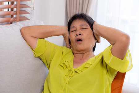 Senior asian woman pain and headache sitting on sofa in living room at home, elderly sitting on couch depression and emotion with stress and migraine, frustration and worried, unhappy and exhaustion.の写真素材