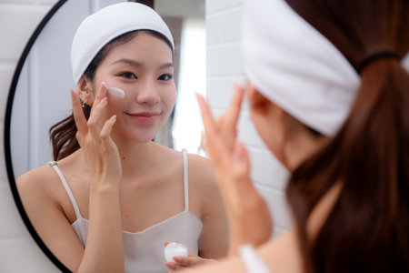 Beautiful young asian woman applying cream or lotion for rejuvenation skin on face while looking mirror in bathroom, woman applying moisturizer for treatment wrinkles on facial, beauty and skincare.の写真素材