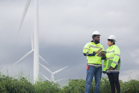 Two engineer check and examining wind turbines in wind farm, renewable energy and teamwork in a sustainable environment, inspection power electric with document and tablet, industry and eco.の写真素材