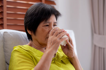 Happiness senior woman sitting on sofa and holding a coffee cup drinking looking out a window in living room at home, elderly drinking beverage for relax, lifestyle and cozy, domestic life.の写真素材