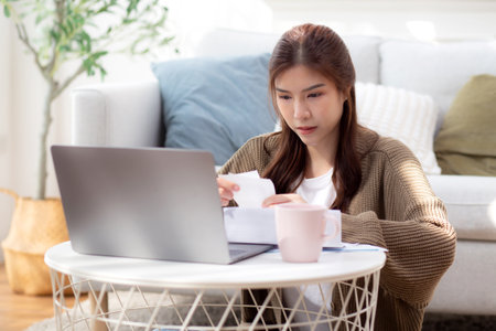 Young asian woman calculates bills finance and cost in living room at home, woman looking expense with burden or investment on paperwork, statement banking and debt for loan, business and financial.の写真素材