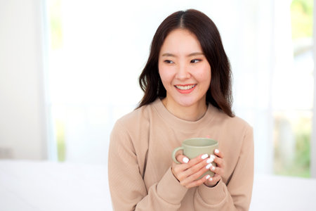 Young asian woman holding and drinking coffee in morning in bedroom at home for relaxation, woman drinking beverage with pleasure and freshness for health, relax and cozy, lifestyle concept.の写真素材