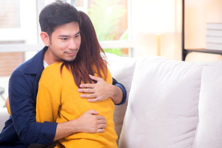 Young asian couple hug sitting on sofa with affectionate in living room at home, husband and wife with gentle and relation with cozy and love while embrace, bonding and comfort, couple and lifestyle.の写真素材