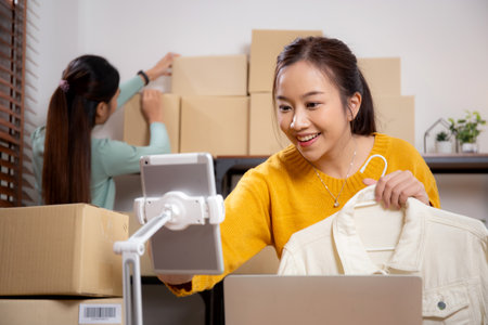 Two young asian woman live online clothing store while showing clothing with tablet and laptop on social at home, entrepreneur streaming for selling online clothing and presenting, online seller.の写真素材