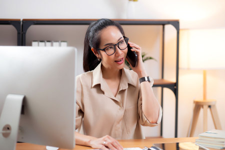 Young asian business woman using desktop computer while talking with smart phone in home office, businesswoman working with desktop computer while speaking with telephone, business or consulting.の写真素材