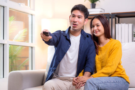 Happy young asian couple sitting on cozy sofa watching TV together and enjoying a relaxation in living room at home, relationship and comfortable of couple during watching television on couch.の写真素材