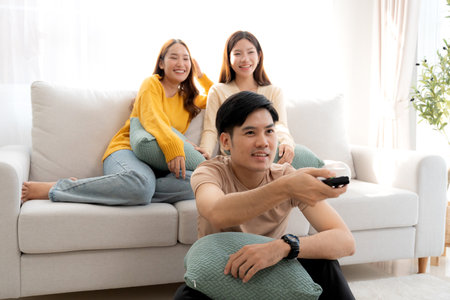 Group young asian friends watching TV together in a cozy living room at home with happy, laughing and enjoying sitting on sofa for watching television with relax, lifestyle and entertainment.の写真素材
