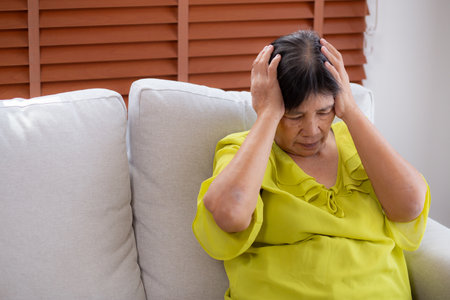 Senior asian woman pain and headache sitting on sofa in living room at home, elderly sitting on couch depression and emotion with stress and migraine, frustration and worried, unhappy and exhaustion.の写真素材